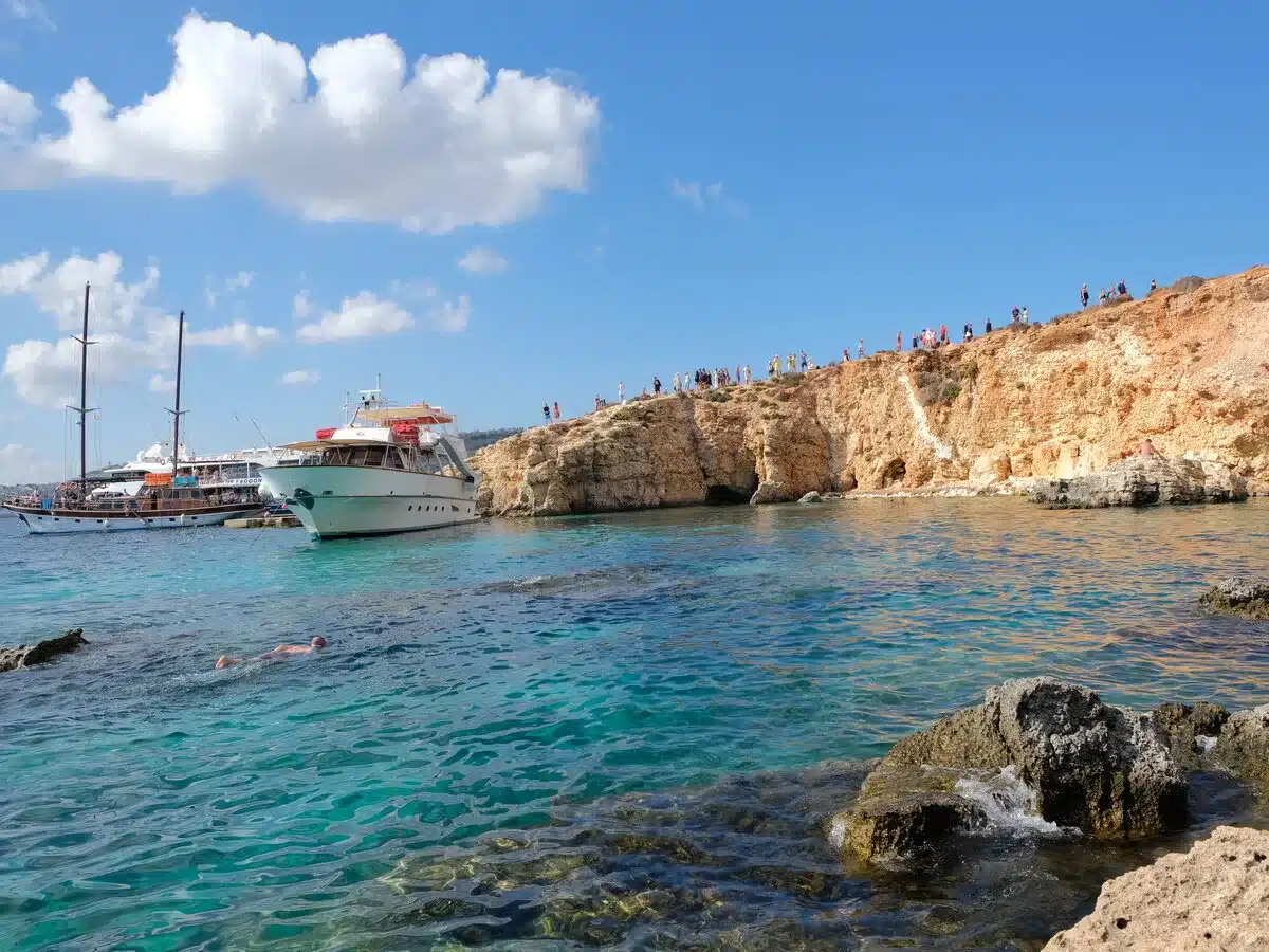 Swimmers and tour boats enjoy the crystal-clear waters of the Blue Lagoon in Malta. A white tour boat dominates the foreground while visitors crowd the sun-kissed, rocky cliffs in the background, under a sky dotted with fluffy clouds.