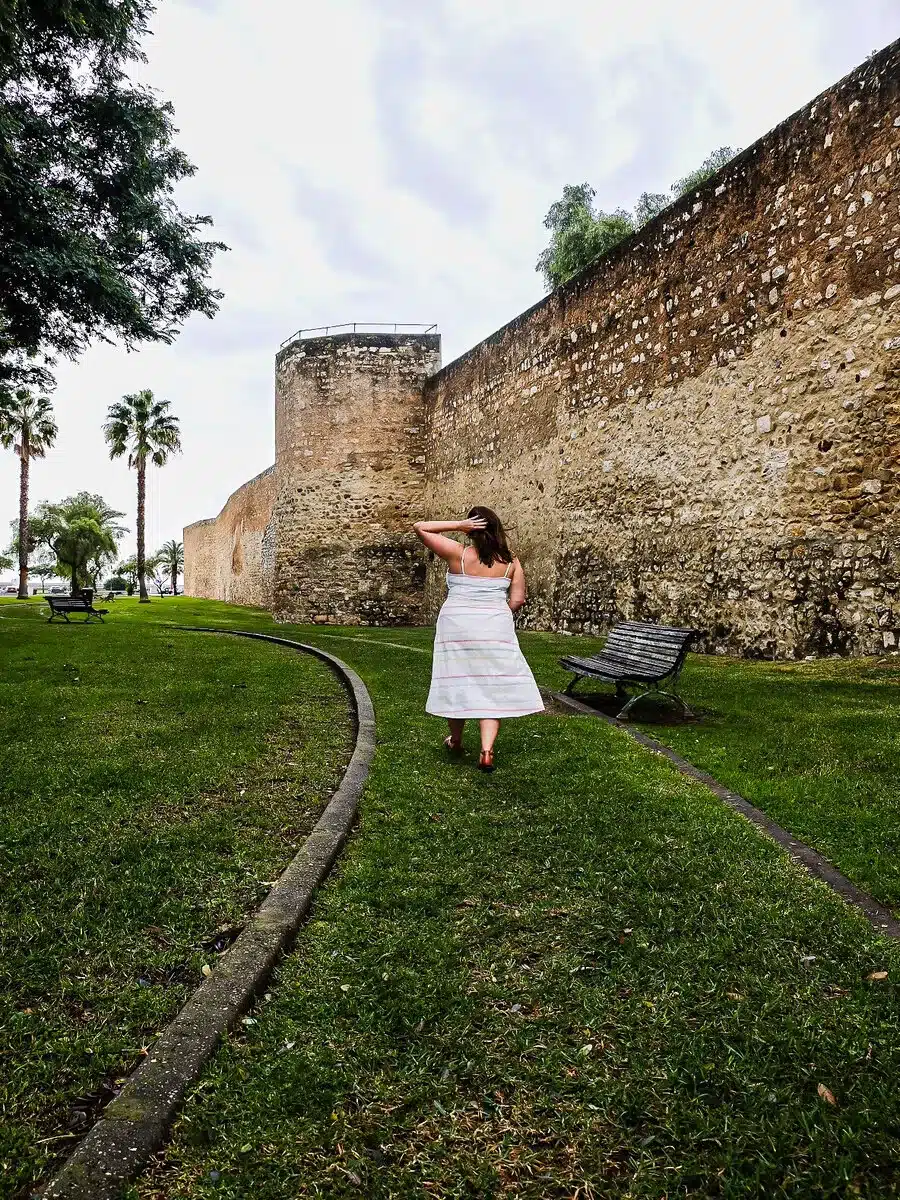 Adventurous solo traveler exploring the historic walls of Faro, surrounded by lush greenery, under the overcast sky, evoking a sense of discovery and solitude.