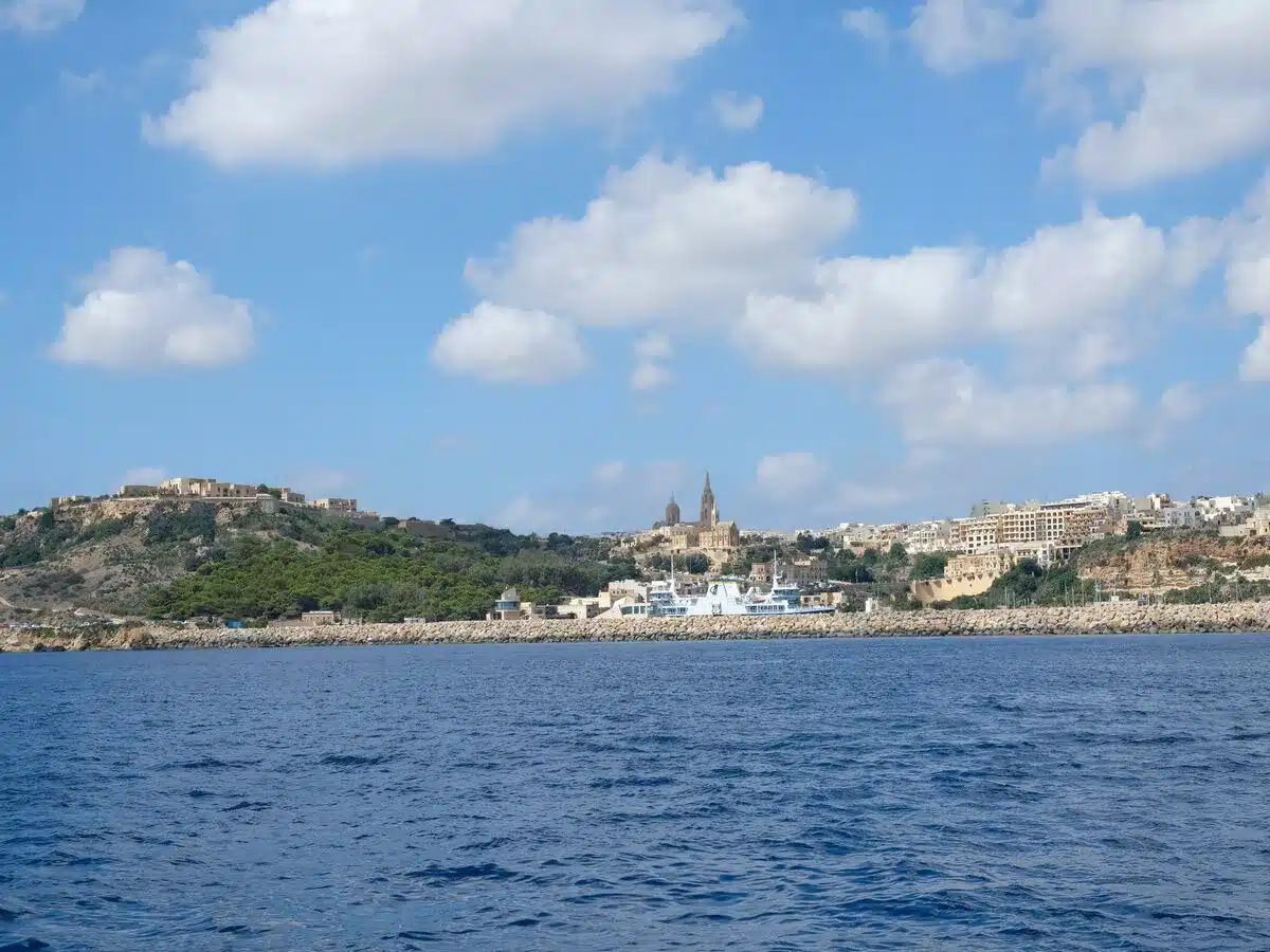 The scenic coastline of Gozo, Malta, viewed from the sea, with historic buildings perched atop cliffs overlooking the deep blue water. The skyline is accented by the imposing Citadel and the baroque steeples of the town's churches, set against a backdrop of a clear sky with fluffy clouds.