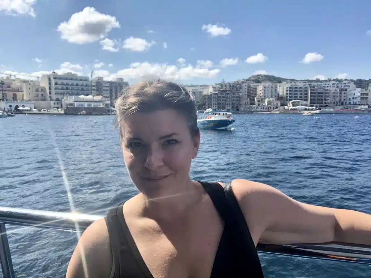A woman enjoys a private boat tour in Malta, resting her arm on the rail with the Mediterranean Sea behind her. The backdrop features the charming architecture of a Maltese town and boats dotting the vibrant harbor under a clear sky with a few scattered clouds.