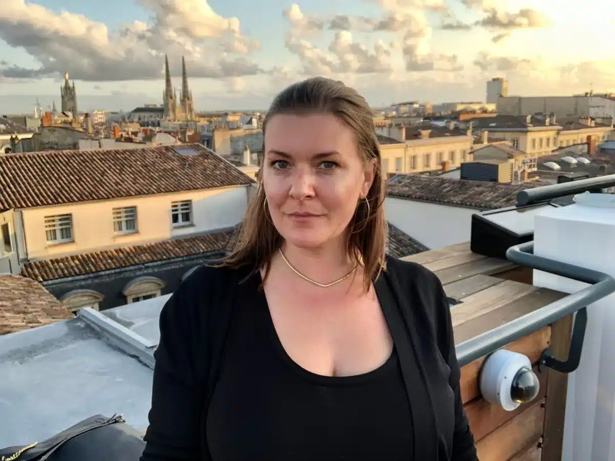 A portrait of a solo female traveler with a scenic city skyline in the background during what appears to be the golden hour. She is wearing a black top and a light cardigan, with a subtle smile, and her hair is swept to one side.