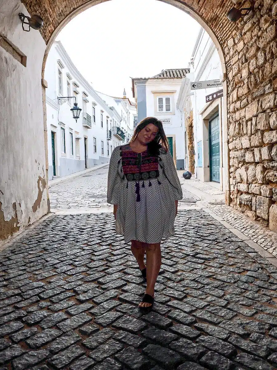 A contented traveler stands on the cobblestone streets of Faro, Portugal, framed by a stone archway and surrounded by traditional white buildings with terracotta roofs.