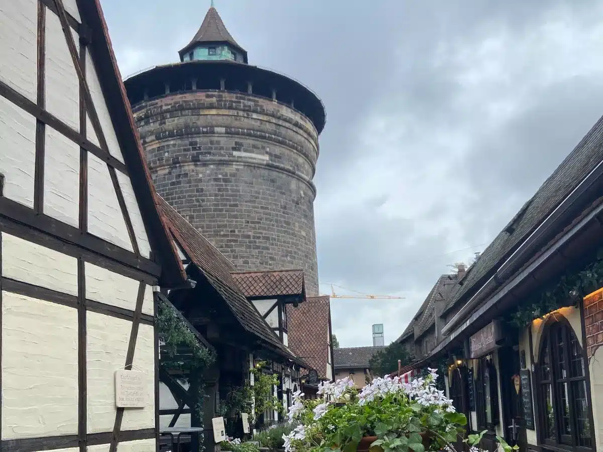 A charming view of Nuremberg's famous Hangman's Bridge, featuring half-timbered structures and the imposing Sinwell Tower. Flowers and greenery add a touch of life to the historic site, inviting visitors to discover the tales of the past.